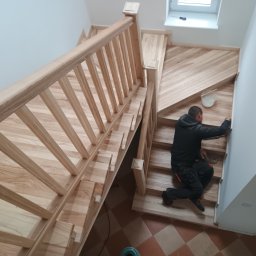 Dom drew - Montaż drewnianych schodów z balustradą przez stolarza. Widok z góry na jasne drewno i pracownika w trakcie wykończenia.