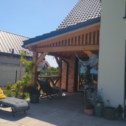 Dom drew - Zadaszony taras z drewnianą konstrukcją, leżanką i roślinami w donicach. Widoczny fragment domu z cegły i jasną elewacją. Oświetlenie w postaci żarówek.