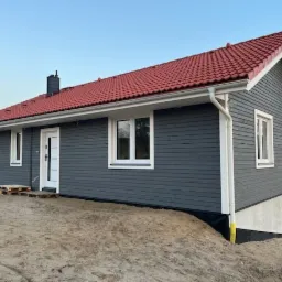 Szary dom szkieletowy z czerwonym dachem, widok z boku. Białe okna i drzwi kontrastują z ciemną elewacją. Teren wok&oacute;ł domu jeszcze niezagospodarowany.