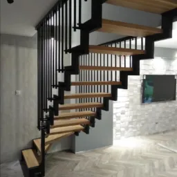 Nowoczesne schody z drewnianymi stopniami i czarną metalową balustradą. Schody spiralne, z widocznym oświetleniem i teksturą ściany w tle.
