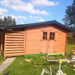 Drewniana altana z dwuskrzydłowymi drzwiami w ogrodzie, z widocznymi narzędziami budowlanymi i elementami konstrukcyjnymi. Słoneczny dzień.