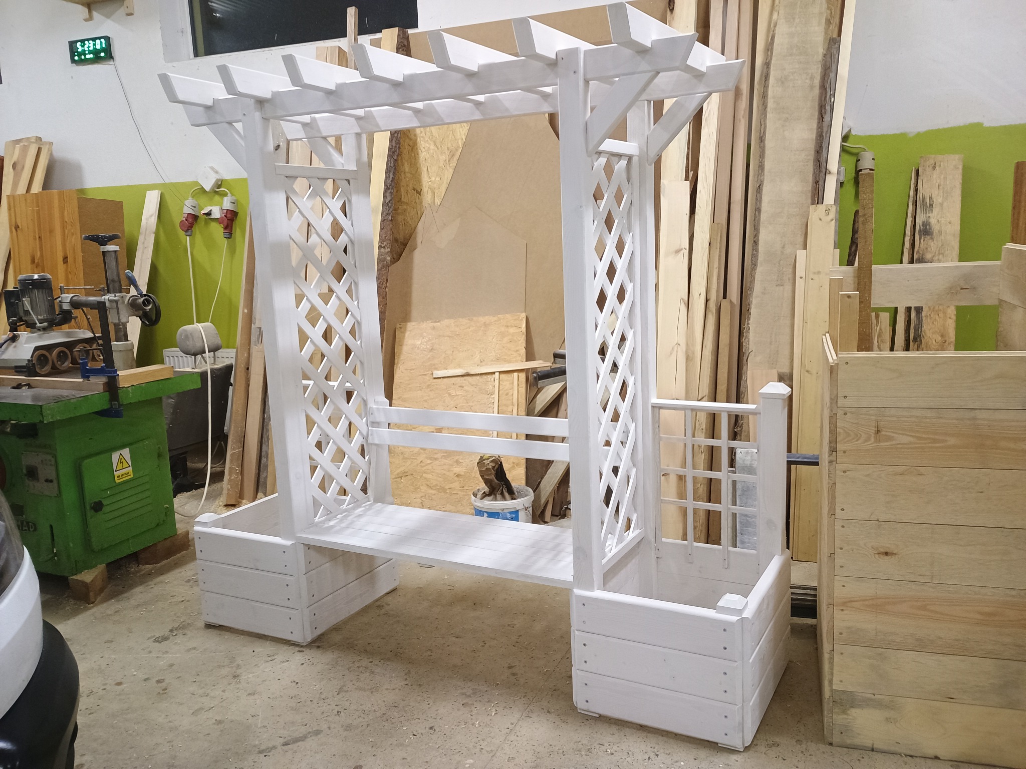 Biała drewniana konstrukcja z kratownicami i siedziskiem, przypominająca małą altanę z donicami, stoi w warsztacie stolarskim, obok maszyny do obróbki drewna i stosu desek.