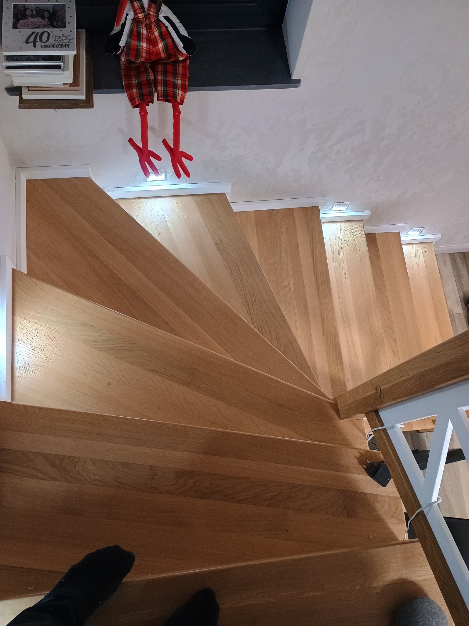 Widok z góry na jasne, drewniane schody z podświetleniem LED w stopniach, u góry dekoracja w postaci pluszowych nóg w czerwono-czarną kratę, na dole widoczne stopy w czarnych skarpetach.