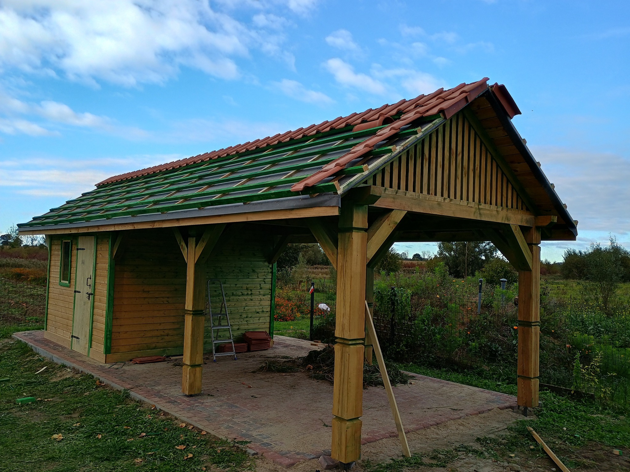Drewniana konstrukcja z częściowo ułożoną czerwoną dachówką, widoczna z zewnątrz, z elementami szalunku dachowego w kolorze zielonym, obok mały domek gospodarczy z zielonymi obramowaniami, teren...