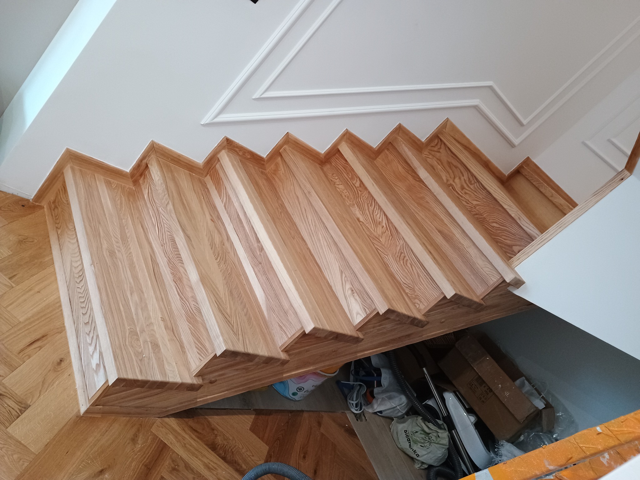 Widok z góry na drewniane schody z jasnego drewna, z widoczną strukturą usłojenia, zamontowane w nowoczesnym wnętrzu z parkietem w jodełkę i białym sufitem z dekoracyjną sztukaterią. Pod schodami...