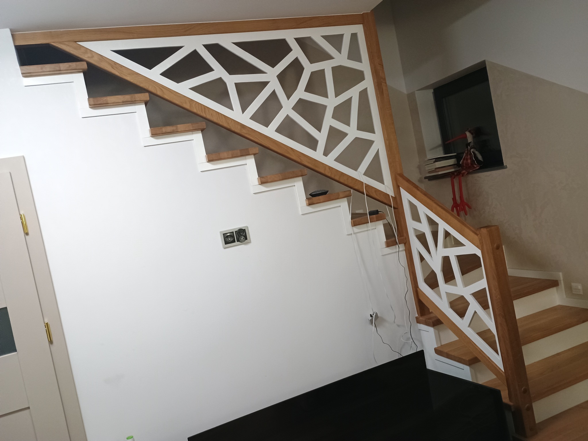 Białe schody z drewnianymi stopniami i geometryczną balustradą z drewna i bieli, widok z dołu, w tle okno z dekoracją.