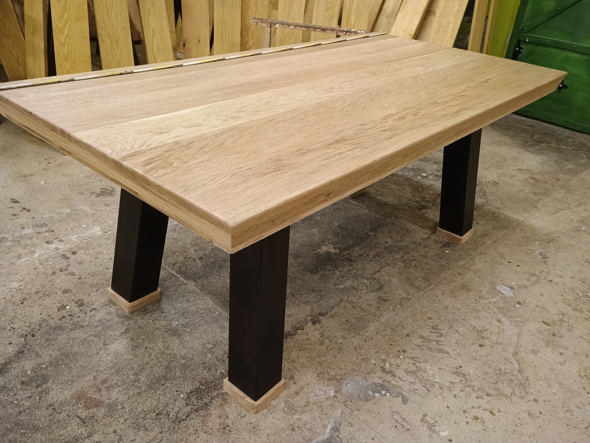 Stół z jasnego drewna z czarnymi, kwadratowymi nogami, ustawiony na betonowej podłodze, z drewnianymi elementami w tle. Na blacie leży miarka.