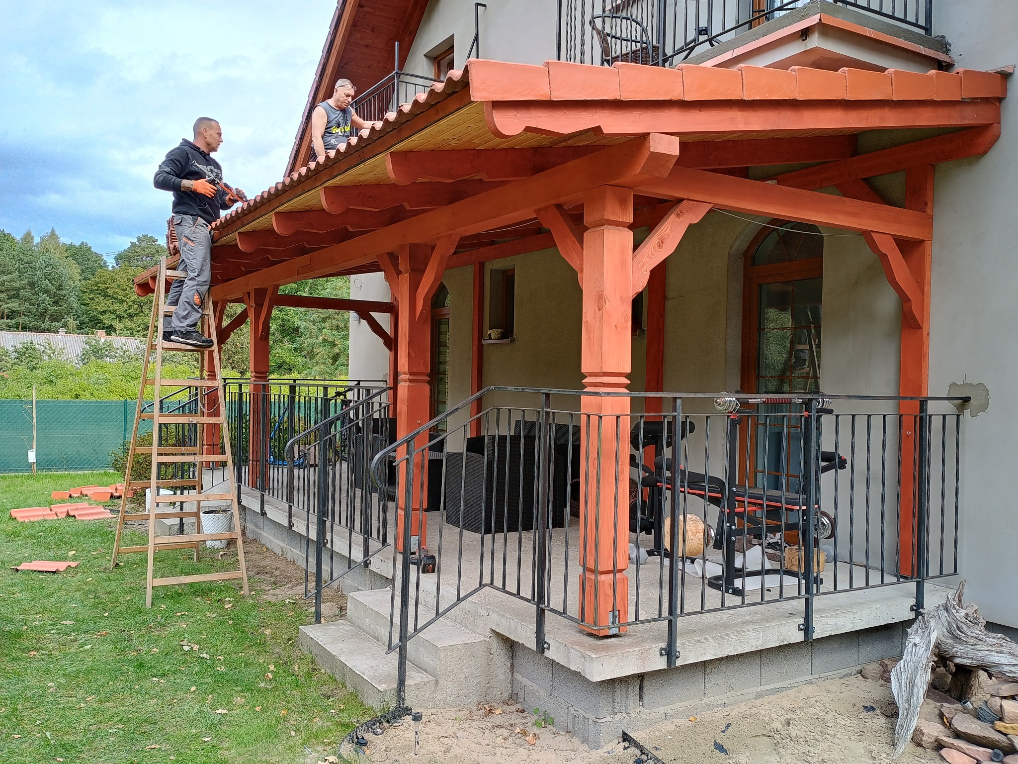 Montaż zadaszenia tarasu z drewnianą konstrukcją i czerwoną dachówką przez dwóch mężczyzn; jeden na drabinie, drugi na dachu, widok na ogród i dom.
