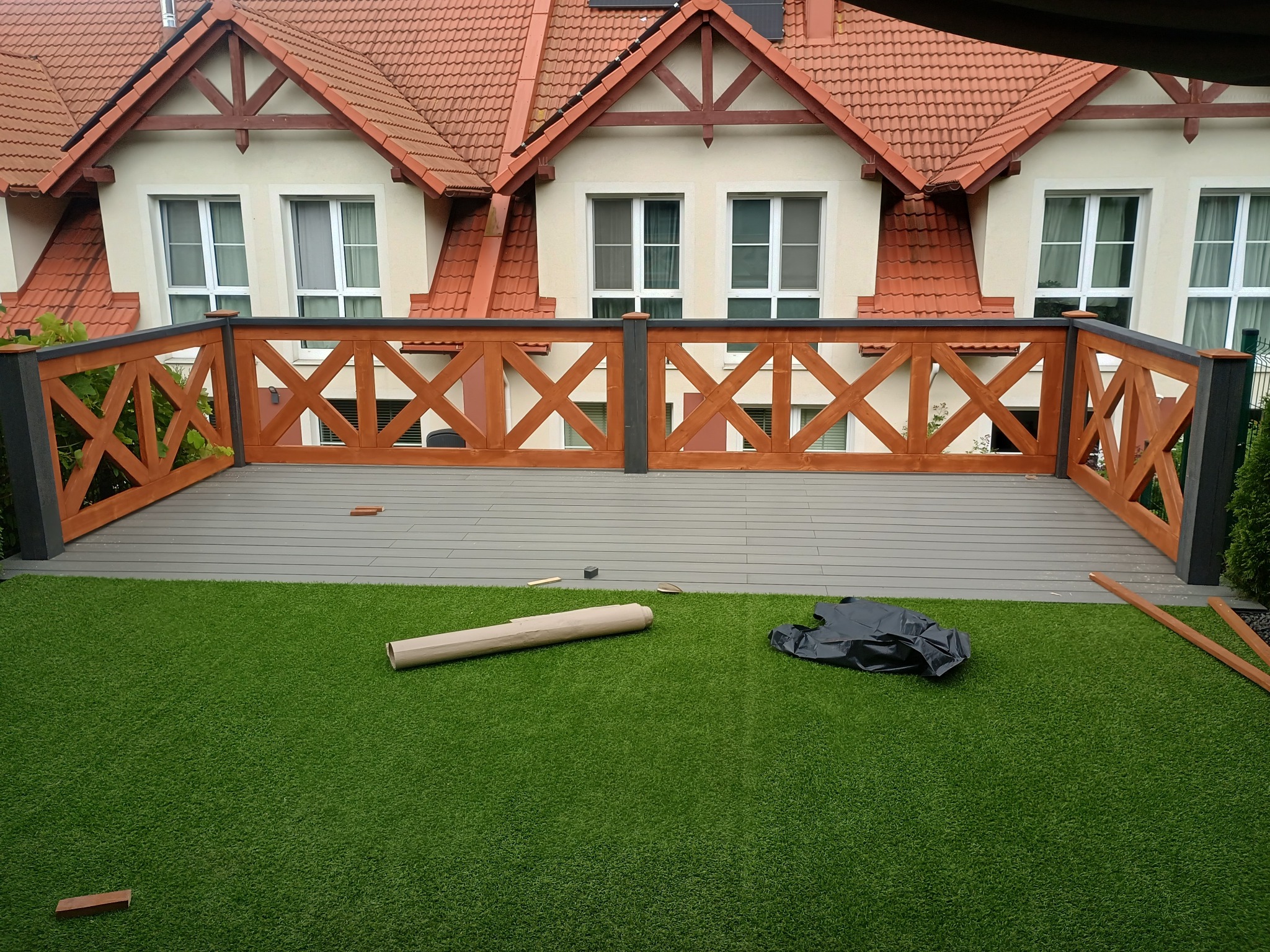 Wykończony taras z drewnianą balustradą w kształcie krzyży, imitacja trawy i elementy montażowe. Tło stanowi budynek z czerwonym dachem.