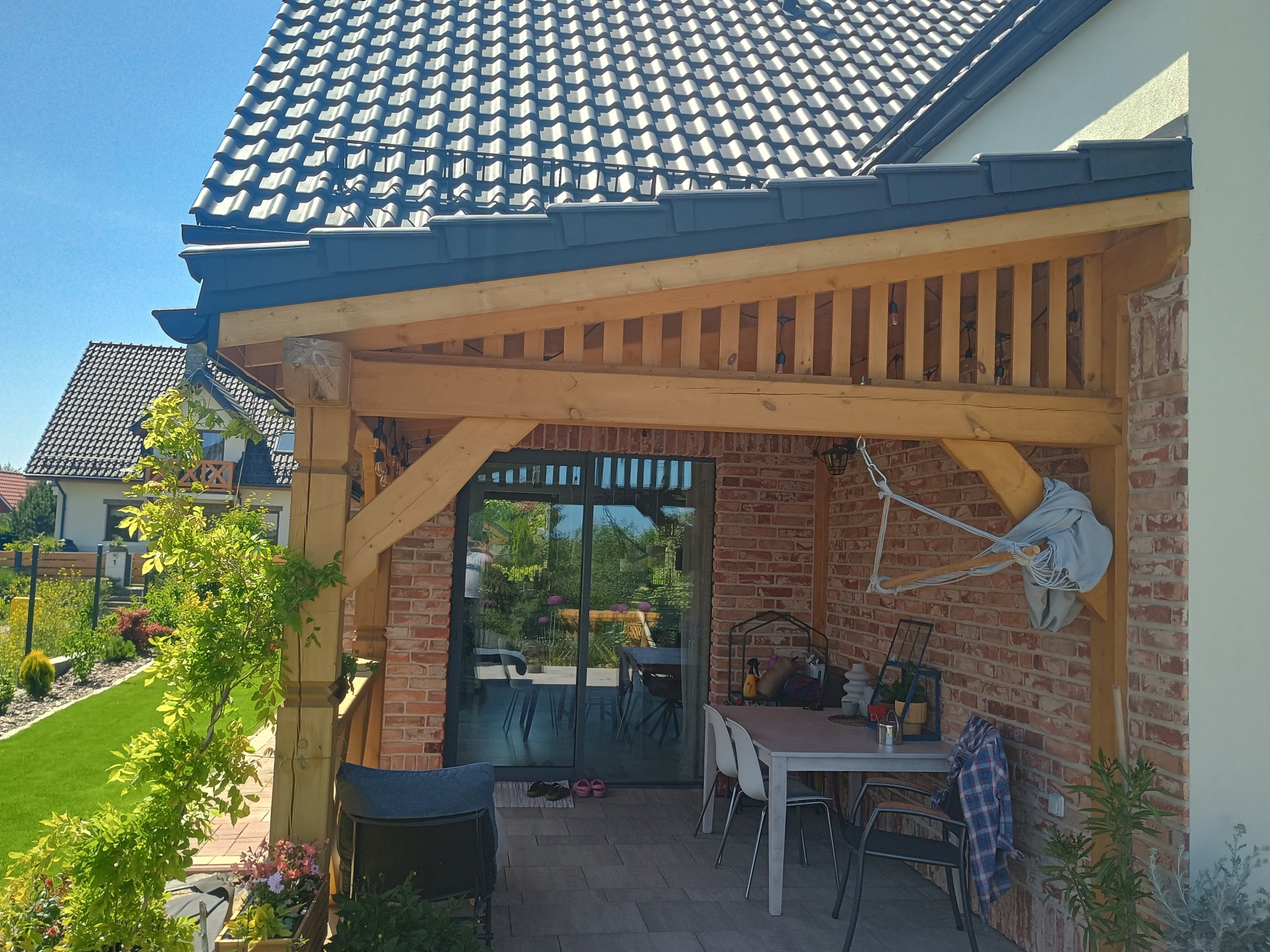 Zadaszony taras z ceglaną ścianą i drewnianą konstrukcją, wyposażony w stół i krzesła, z widokiem na ogród przez przesuwne drzwi.