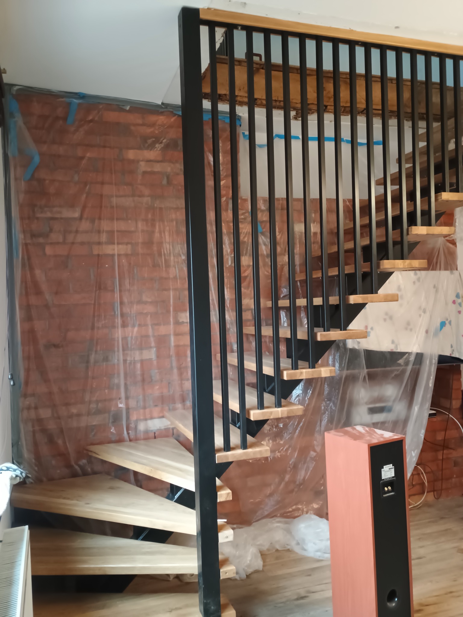 Wnętrze w trakcie wykańczania z drewnianymi schodami i czarną balustradą. Ściany zabezpieczone folią malarską, widoczny głośnik. Ujęcie z dołu, perspektywa żabia.