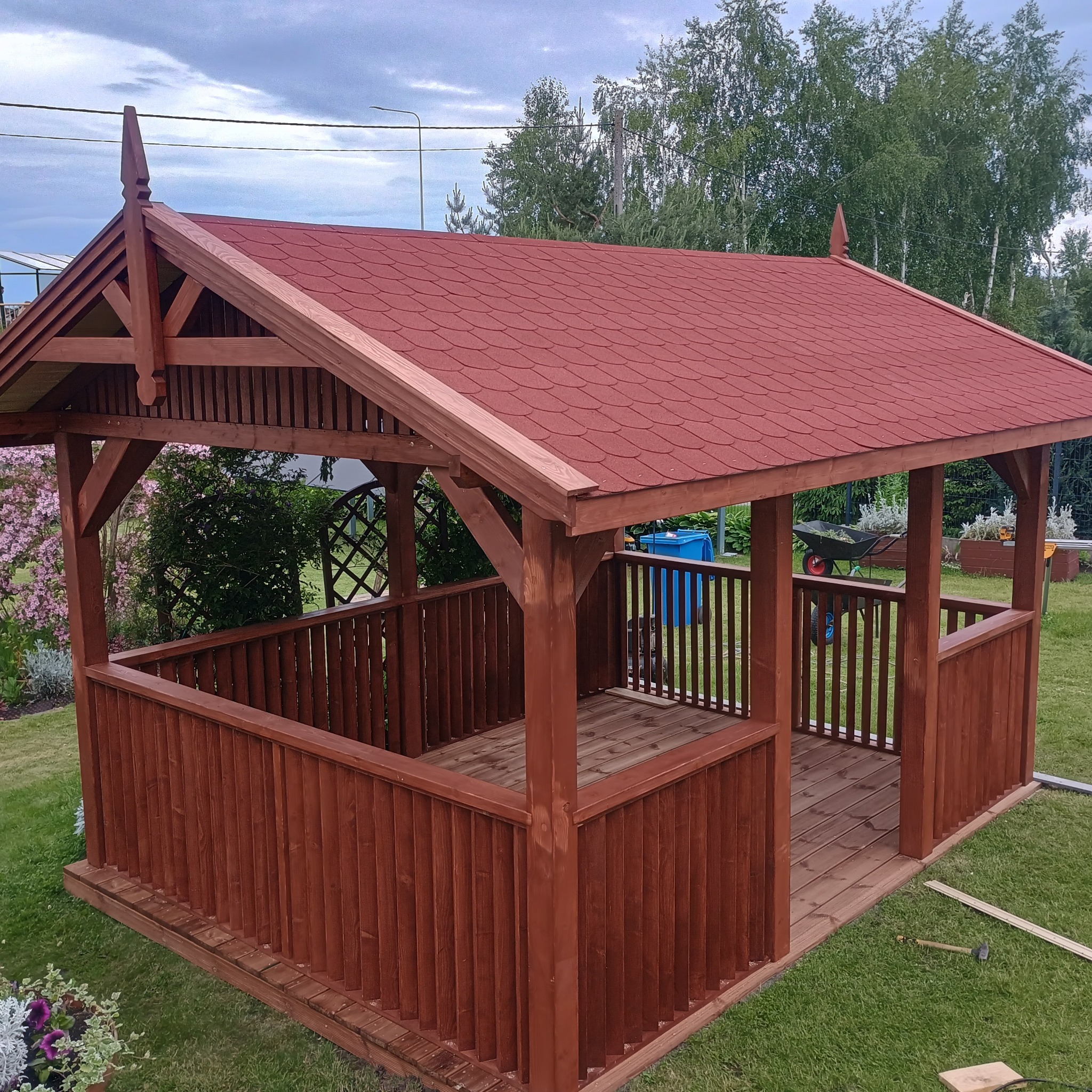 Zadaszona, brązowa konstrukcja z czerwonym dachem na trawniku, otoczona zielenią. Widoczne elementy dekoracyjne na dachu i narzędzia na trawie.