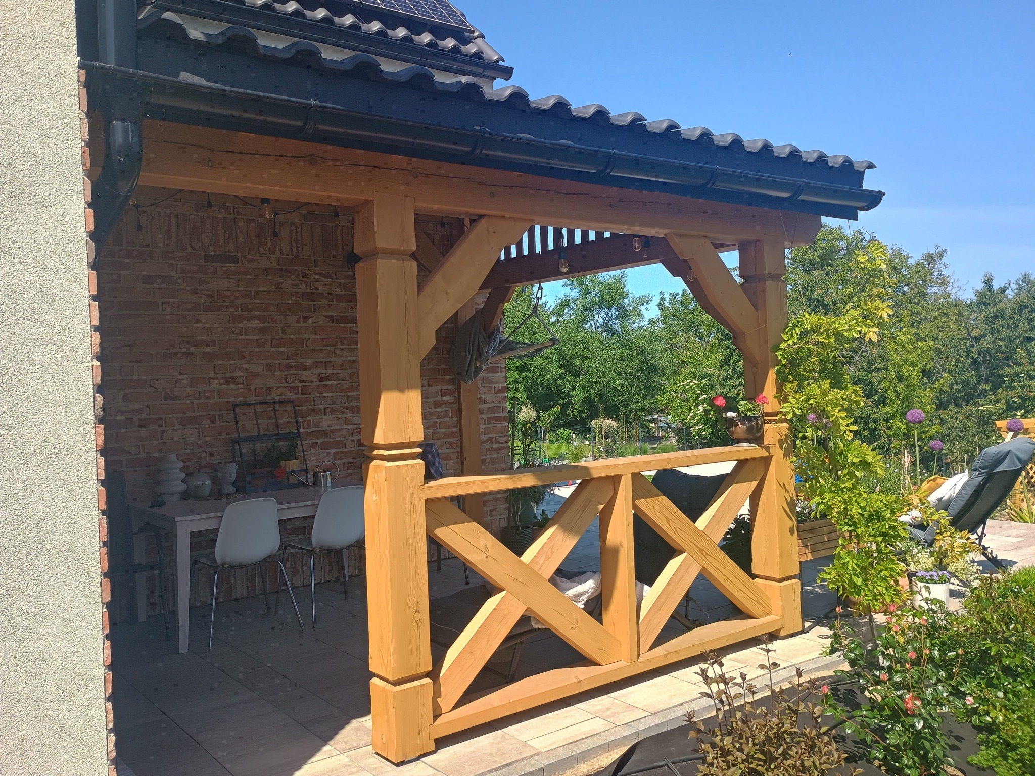 Zadaszony taras z jasnego drewna z ozdobną balustradą krzyżową. W tle ceglana ściana domu i bujna roślinność.