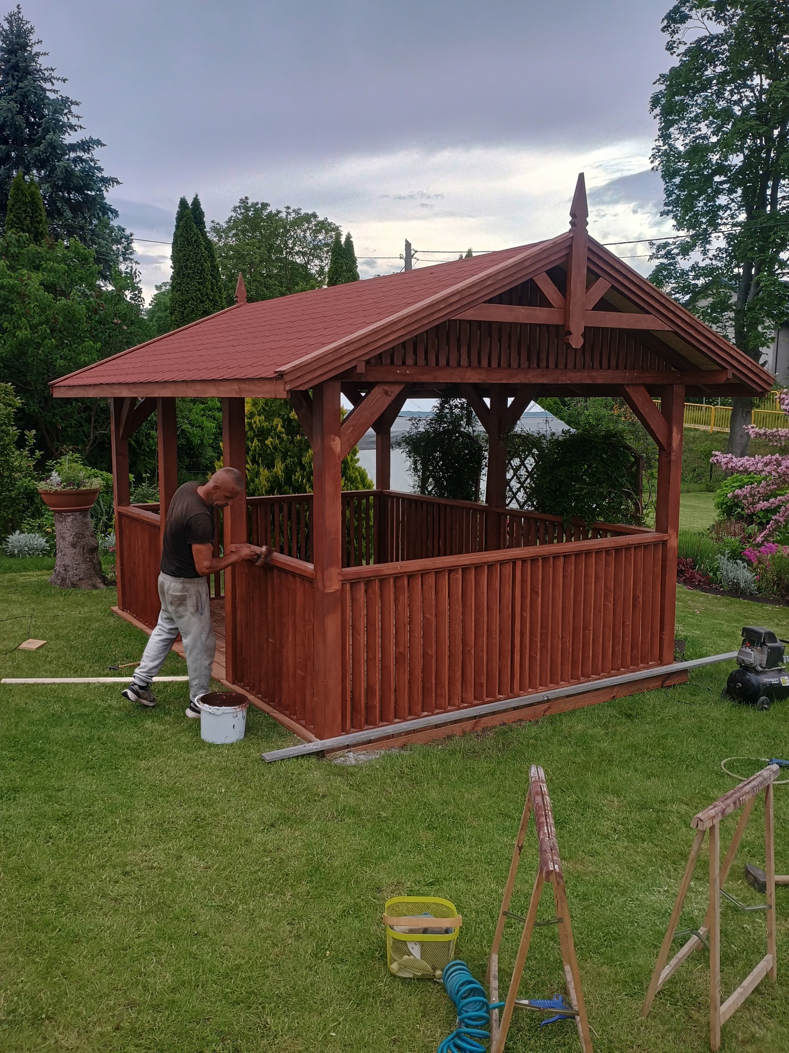 Malowanie drewnianej altany z czerwonym dachem. Mężczyzna maluje balustradę na zielonym trawniku. Widoczne narzędzia i wiadro z farbą.