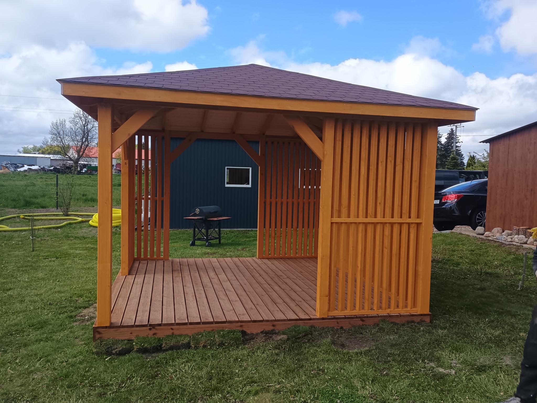 Drewniana altana z brązowym dachem na trawniku, z widocznym grillem i elementami otoczenia. Konstrukcja z jasnego drewna z pionowymi listwami.