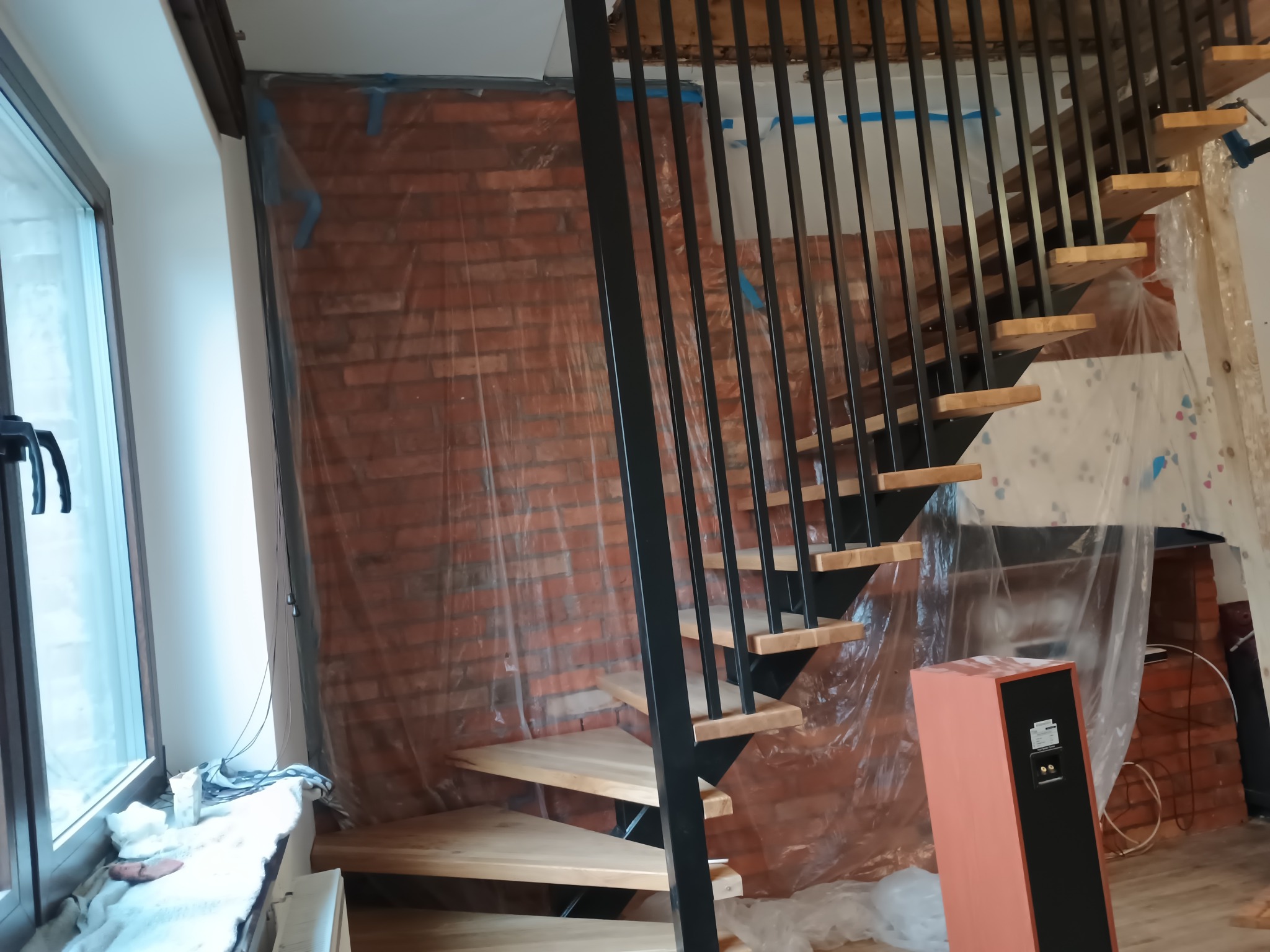 Montaż schodów: surowe drewniane stopnie z czarną metalową balustradą na tle ceglanej ściany zabezpieczonej folią. Widoczne elementy wykończeniowe.