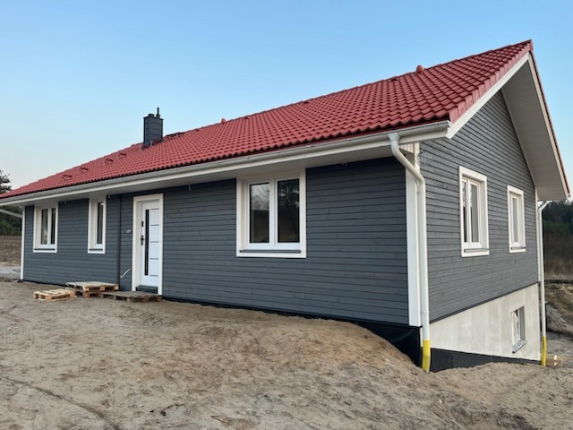 Szary dom szkieletowy z czerwonym dachem, widok z boku. Białe okna i drzwi kontrastują z ciemną elewacją. Teren wokół domu jeszcze niezagospodarowany.