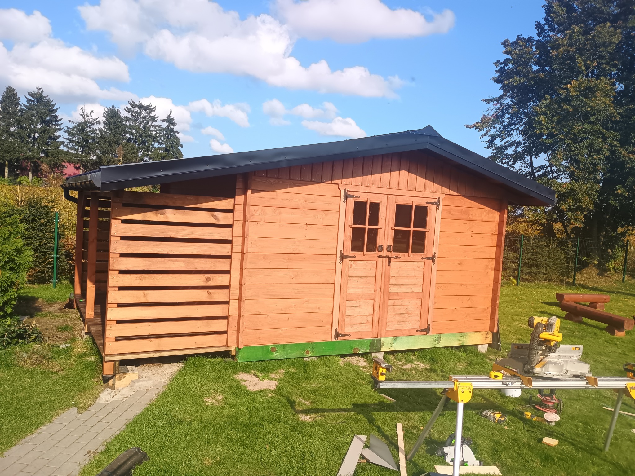Drewniana altana z dwuskrzydłowymi drzwiami w ogrodzie, z widocznymi narzędziami budowlanymi i elementami konstrukcyjnymi. Słoneczny dzień.