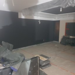 Alek Pisarczyk - Wnętrze w trakcie prac: ściana pomalowana na ciemny grafit, folia ochronna na suficie, materiały budowlane na podłodze z widocznymi liniami poziomowania.