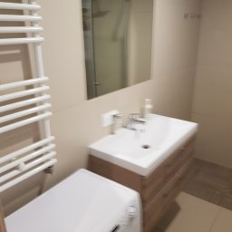 Alek Pisarczyk - Jasna łazienka po remoncie: umywalka z szafką o drewnianym wzorze, biała pralka pod grzejnikiem drabinkowym, lustro i kabina prysznicowa w tle.