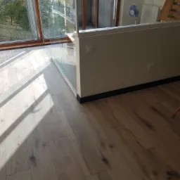 Jasne panele podłogowe w nowym mieszkaniu, oświetlone promieniami słonecznymi wpadającymi przez duże okno z drewnianą ramą, widoczny fragment białej ściany i krzesła.