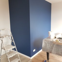 Alek Pisarczyk - Pomieszczenie po malowaniu z granatową ścianą narożną, białe ściany boczne, drabina malarska z resztkami farby, stół zabezpieczony folią i akcesoria malarskie.
