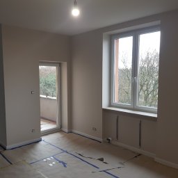 Alek Pisarczyk - Pokój po świeżym malowaniu ścian na beżowo, zabezpieczony folią i taśmą malarską, widoczne okna z białymi ramami i balkon.