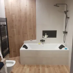 Nowoczesna łazienka z drewnopodobnymi płytkami na podłodze i części ściany, biała wanna z czarnymi zagłówkami, czarna armatura prysznicowa i grzejnik drabinkowy, toaleta z pudełkiem obok.