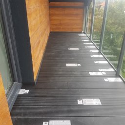 Alek Pisarczyk - Nowo ułożona, ciemnoszara podłoga z desek kompozytowych na balkonie z przeszkloną ścianą, widoczne etykiety producenta na poszczególnych deskach.