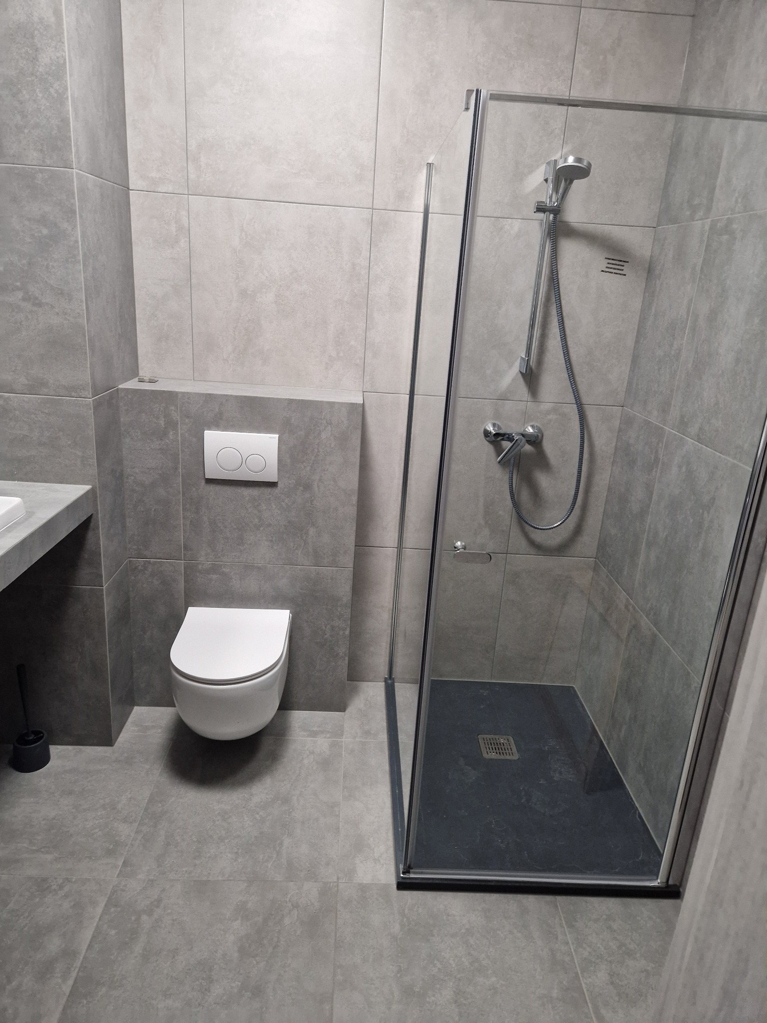 Nowoczesna łazienka w szarościach z kabiną prysznicową, podwieszaną toaletą i umywalką na blacie, wykończona płytkami imitującymi kamień.