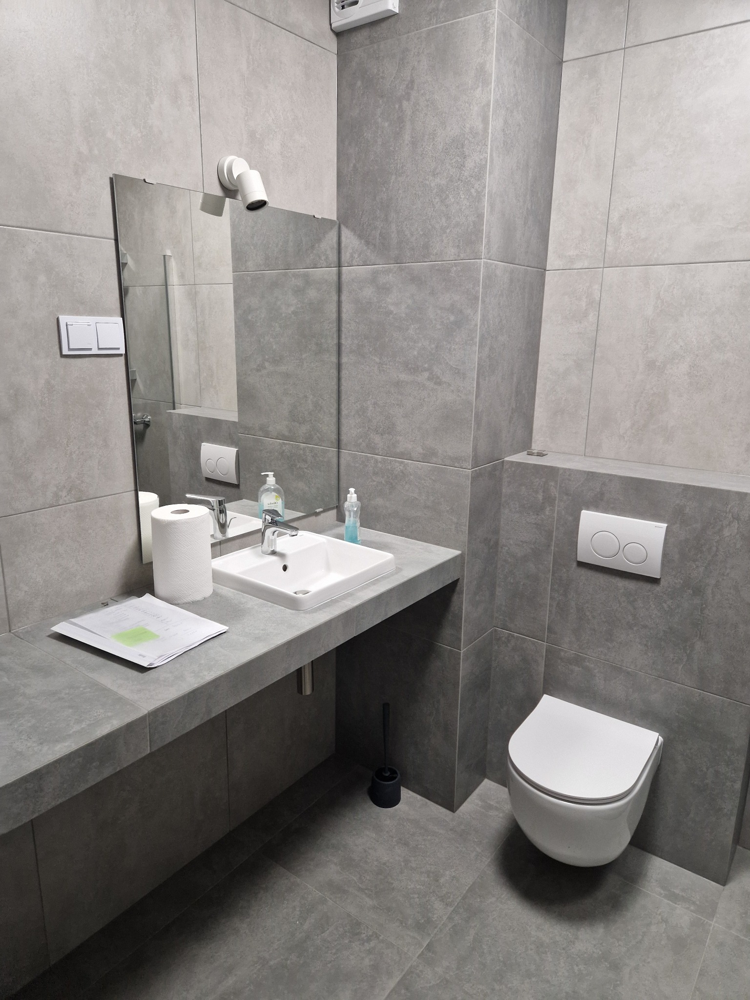 Nowoczesna łazienka w odcieniach szarości z wiszącą toaletą, umywalką nablatową i dużym lustrem, wykończona płytkami ceramicznymi o matowej powierzchni. Na blacie leżą dokumenty i rolka papieru...