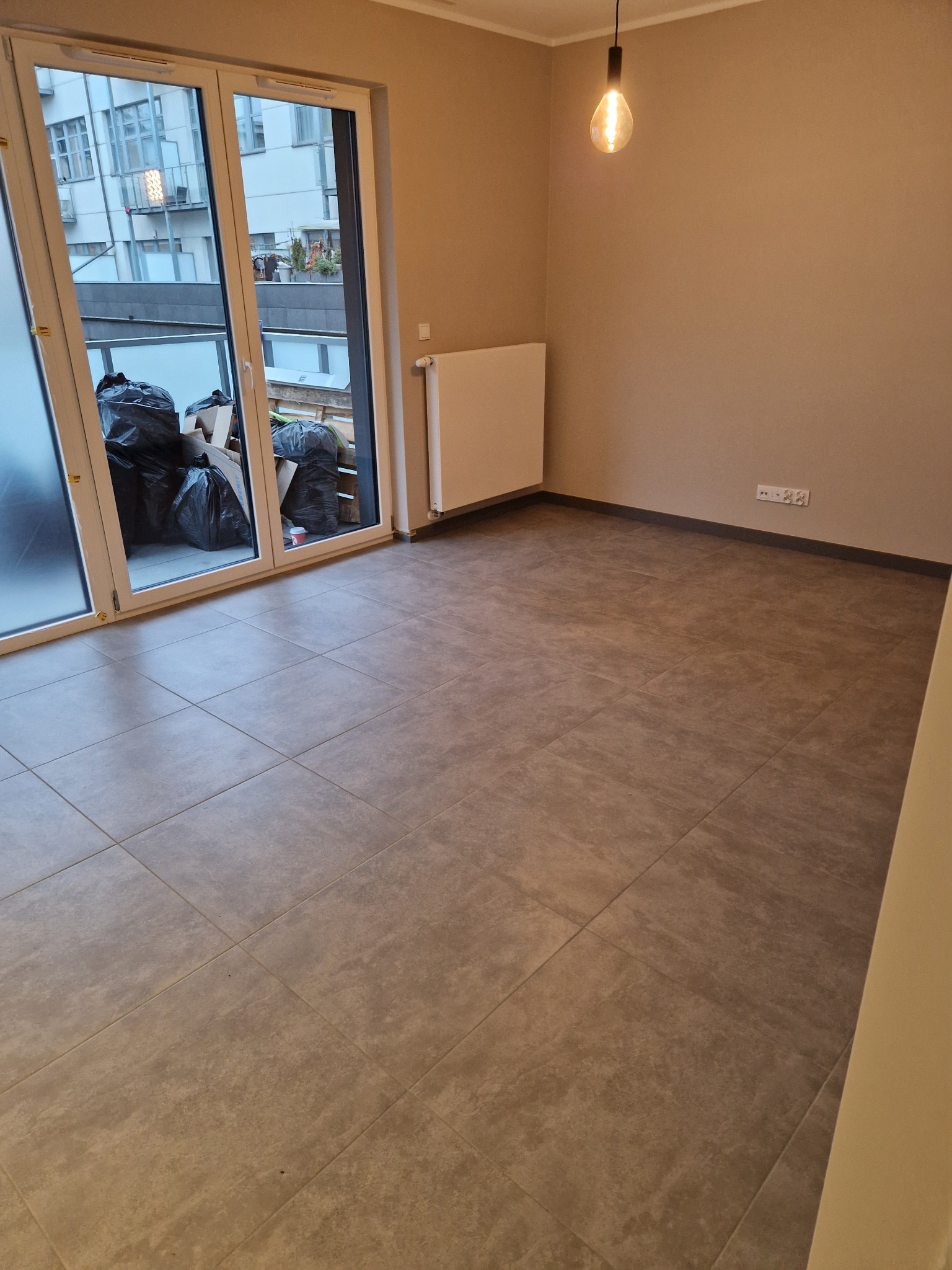 Pokój z szarymi płytkami podłogowymi, grzejnikiem na ścianie, balkonem z widocznymi workami na śmieci i wiszącą żarówką