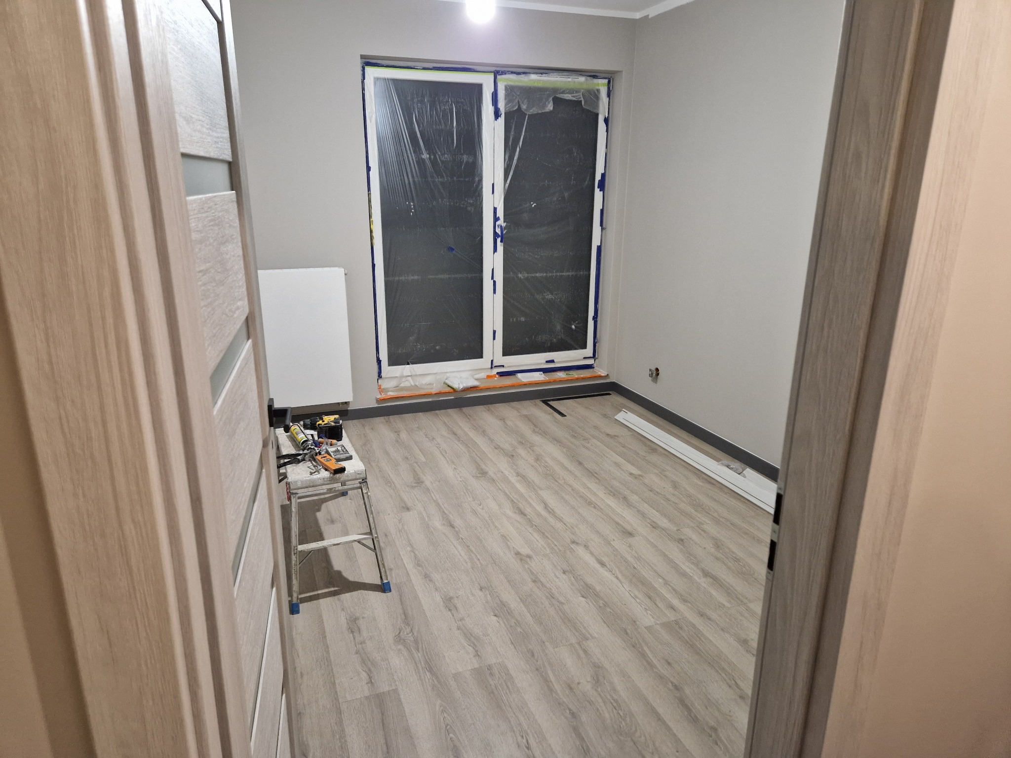 Pokój w trakcie prac wykończeniowych: świeżo położone panele podłogowe w odcieniach szarości, okno zabezpieczone folią malarską, drabina z narzędziami obok drzwi.