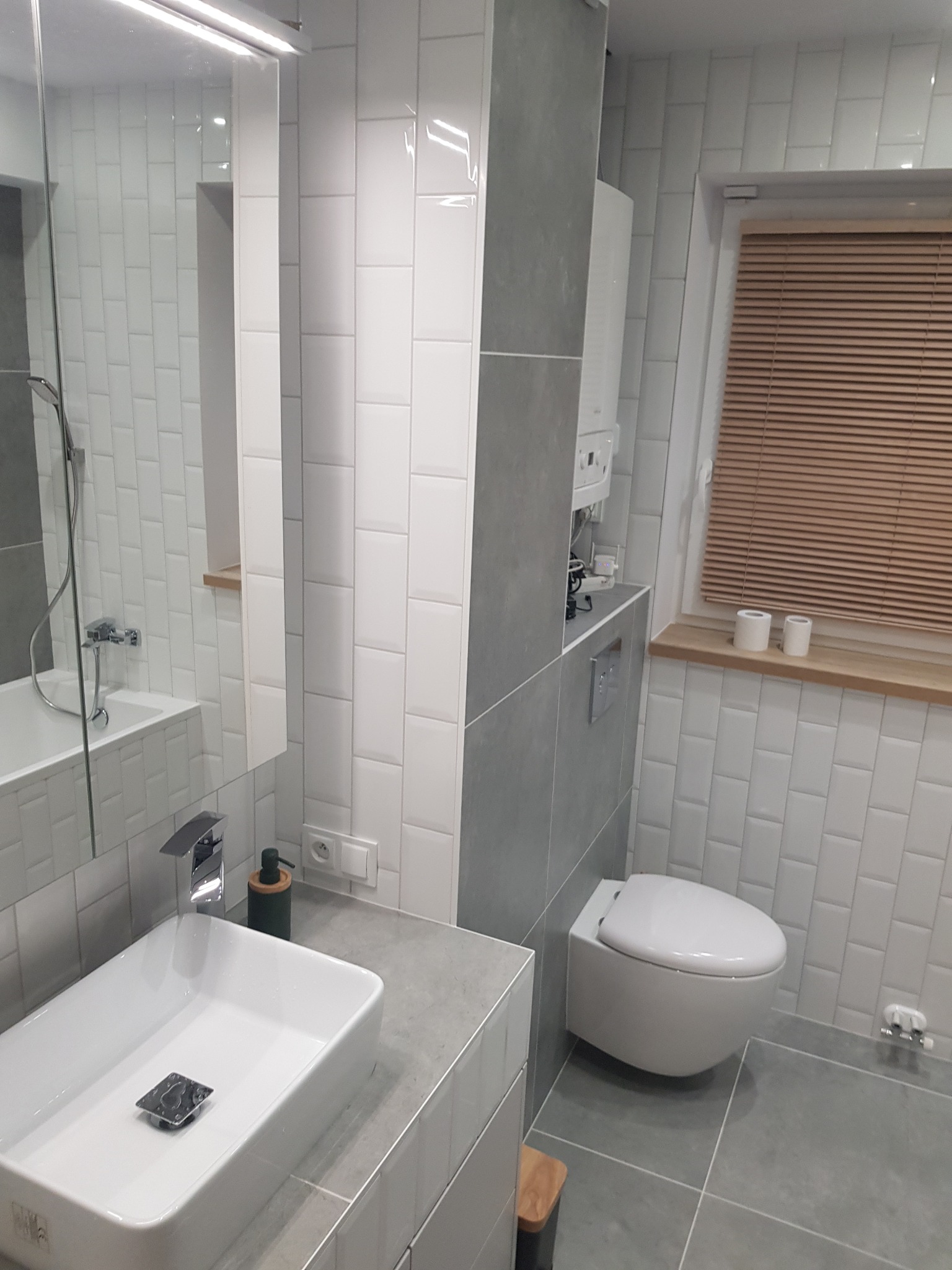 Nowoczesna łazienka z białymi, prostokątnymi płytkami ułożonymi w cegiełkę, szarą zabudową, wiszącą toaletą i drewnianymi żaluzjami w oknie.