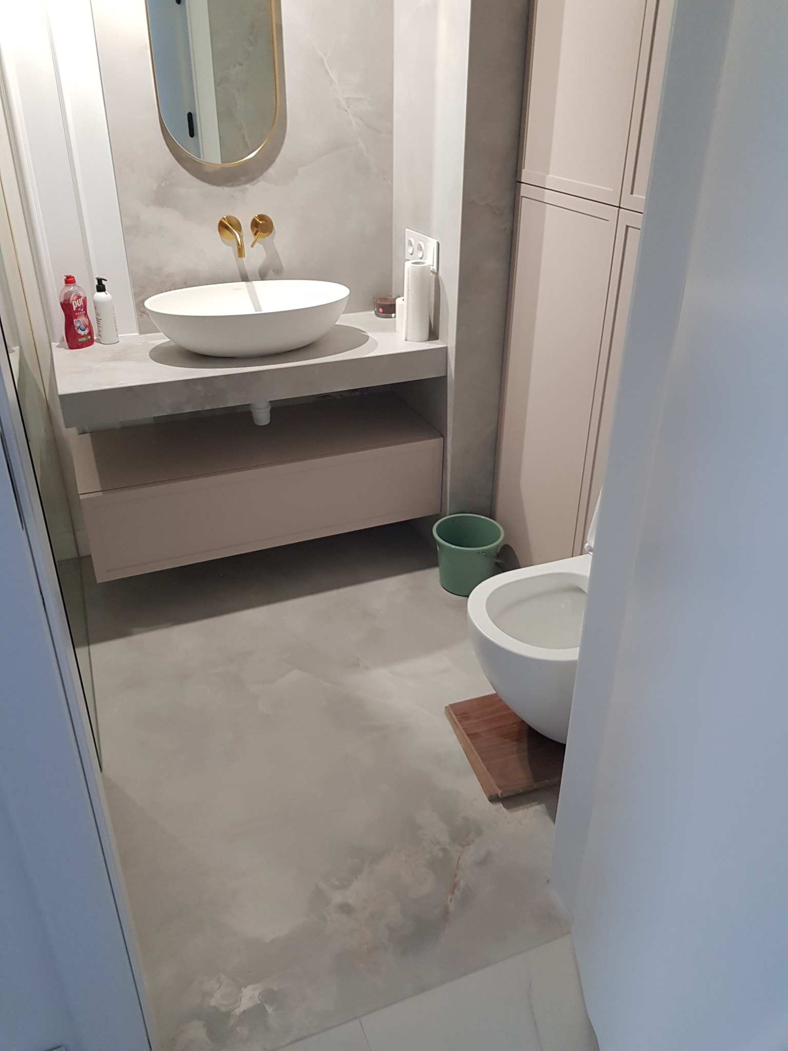 Nowoczesna łazienka z owalną umywalką nablatową, złotą armaturą, lustrem, beżowymi szafkami i szarą podłogą imitującą marmur.