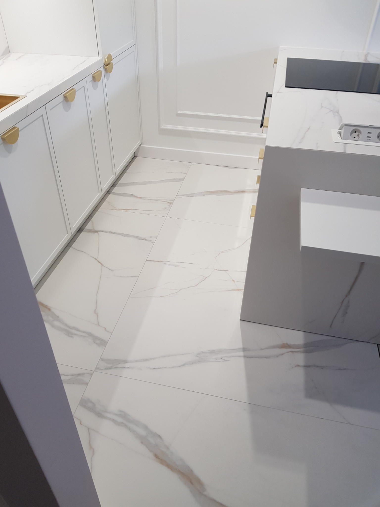 Biała kuchnia z marmurową podłogą i złotymi uchwytami, widok na fragment zabudowy kuchennej, wyspę z wbudowaną płytą i gniazdkami.