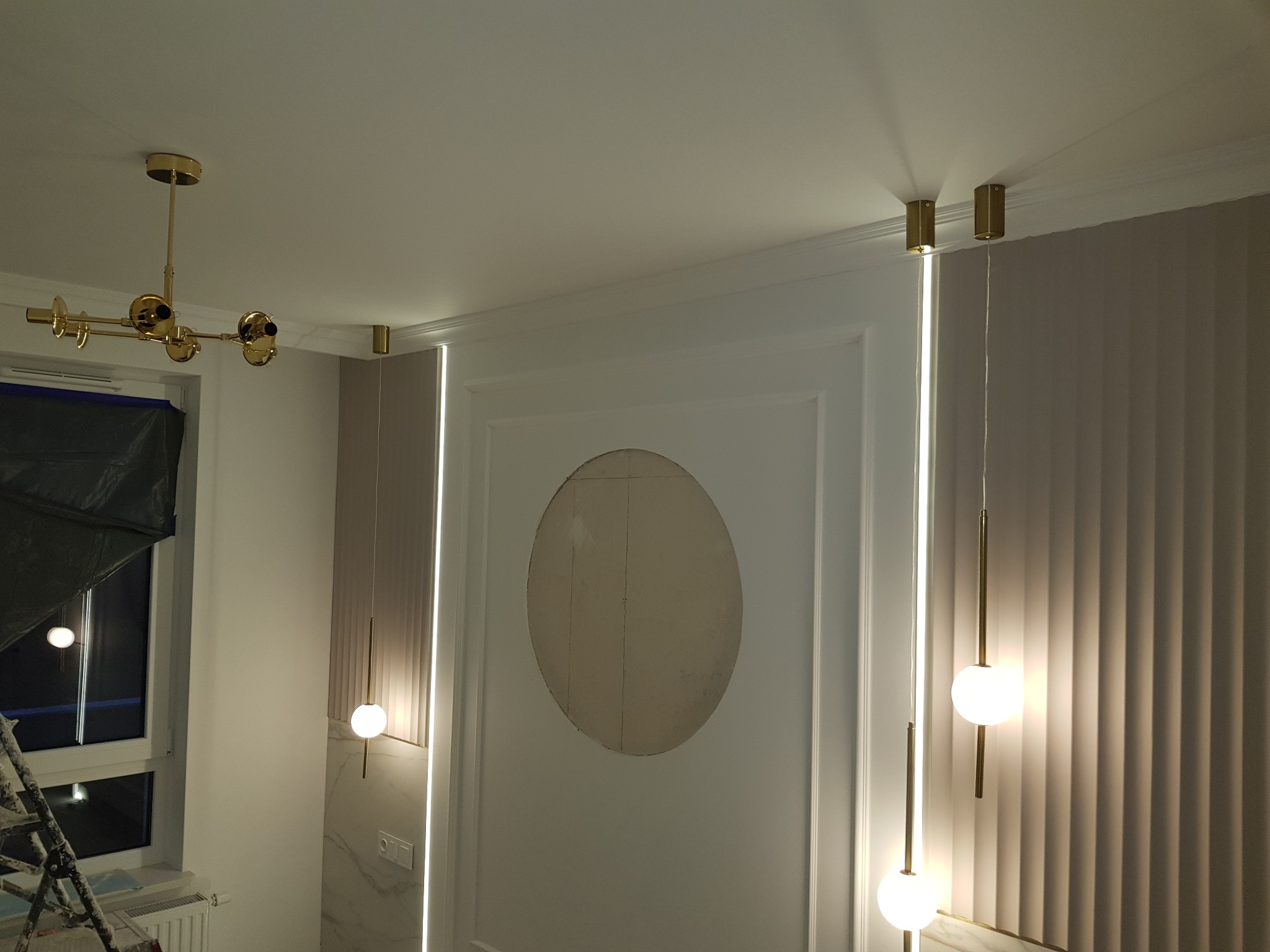 Wnętrze w trakcie prac wykończeniowych z nowoczesnym oświetleniem: złota lampa sufitowa, pionowe lampy kuliste, ściana z dekoracyjną wnęką w kształcie elipsy przygotowana do wykończenia, drabina...