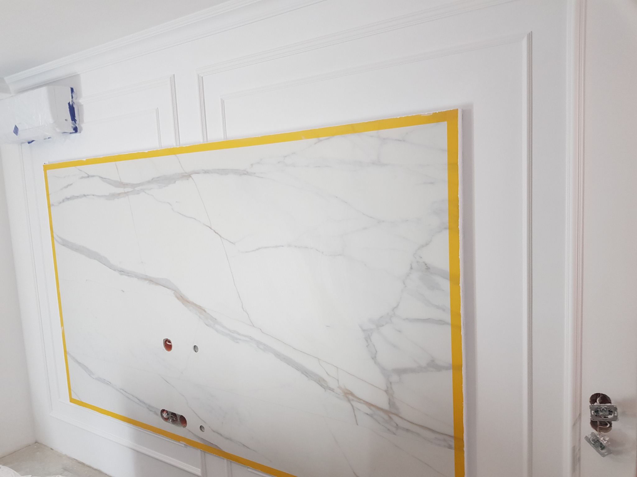 Płyta imitująca marmur z żółtą taśmą malarską, zamontowana na ścianie z ozdobnymi listwami, widoczne puszki elektryczne bez osprzętu i klimatyzator zabezpieczony folią.