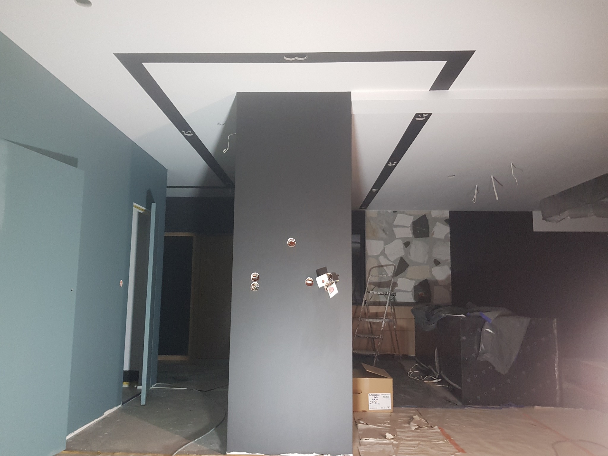 Wnętrze w trakcie prac wykończeniowych: szara kolumna, pomalowana ściana w odcieniach błękitu, sufit z czarnymi dekoracyjnymi pasami, widoczne puszki elektryczne i drabina.