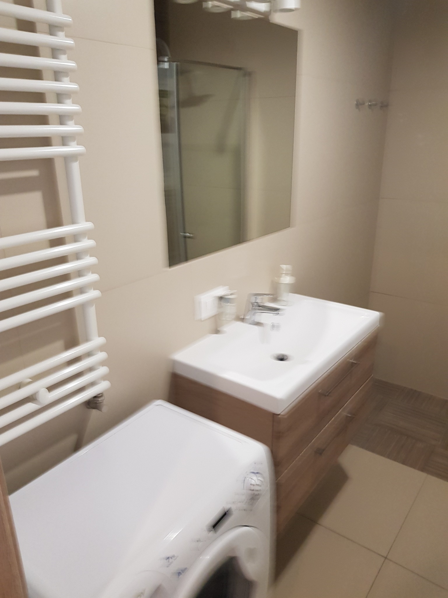 Jasna łazienka po remoncie: umywalka z szafką o drewnianym wzorze, biała pralka pod grzejnikiem drabinkowym, lustro i kabina prysznicowa w tle.