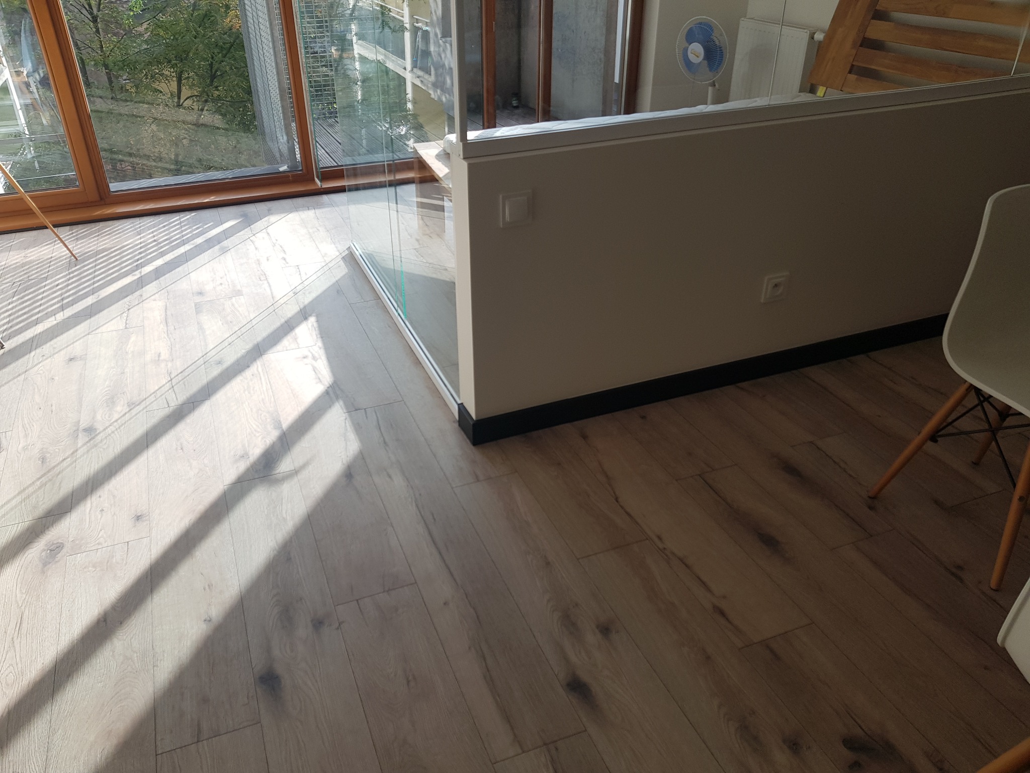 Jasne panele podłogowe w nowym mieszkaniu, oświetlone promieniami słonecznymi wpadającymi przez duże okno z drewnianą ramą, widoczny fragment białej ściany i krzesła.