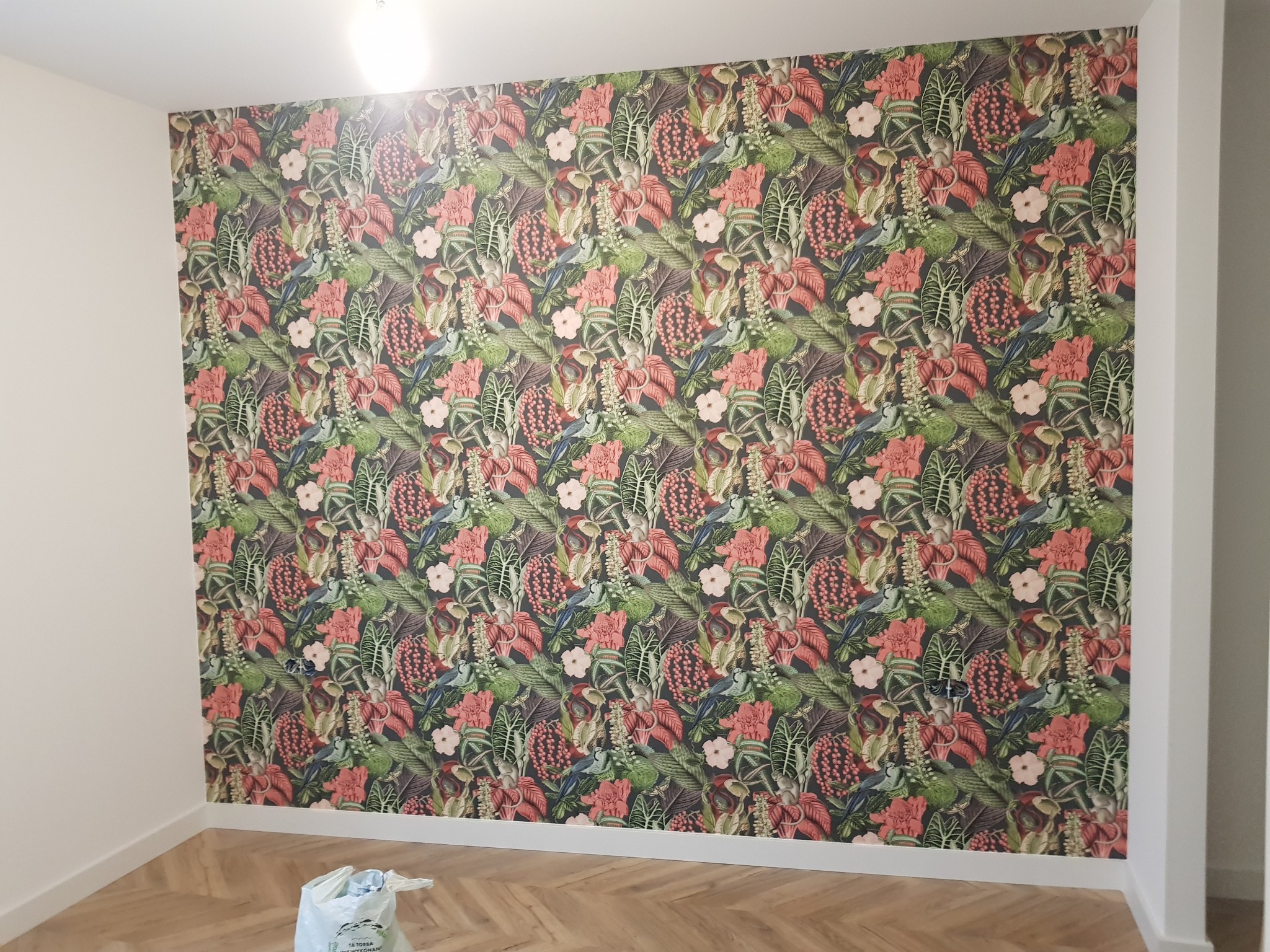 Ściana po zakończonym tapetowaniu w stylu tropikalnym, wzór z motywami roślinnymi i ptakami, parkiet ułożony w jodełkę, widoczny worek z odpadami po pracy.