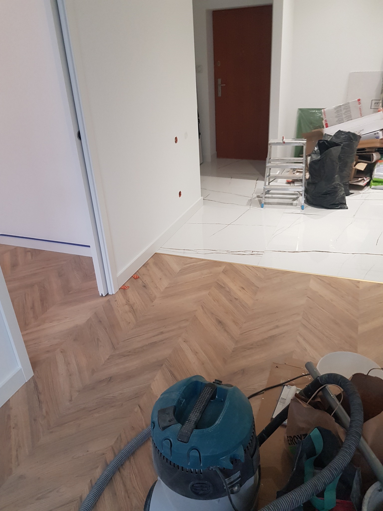 Fragment pokoju z ułożonym parkietem w jodełkę, widoczny odkurzacz budowlany, przejście do pomieszczenia z białymi płytkami podłogowymi i brązowymi drzwiami.