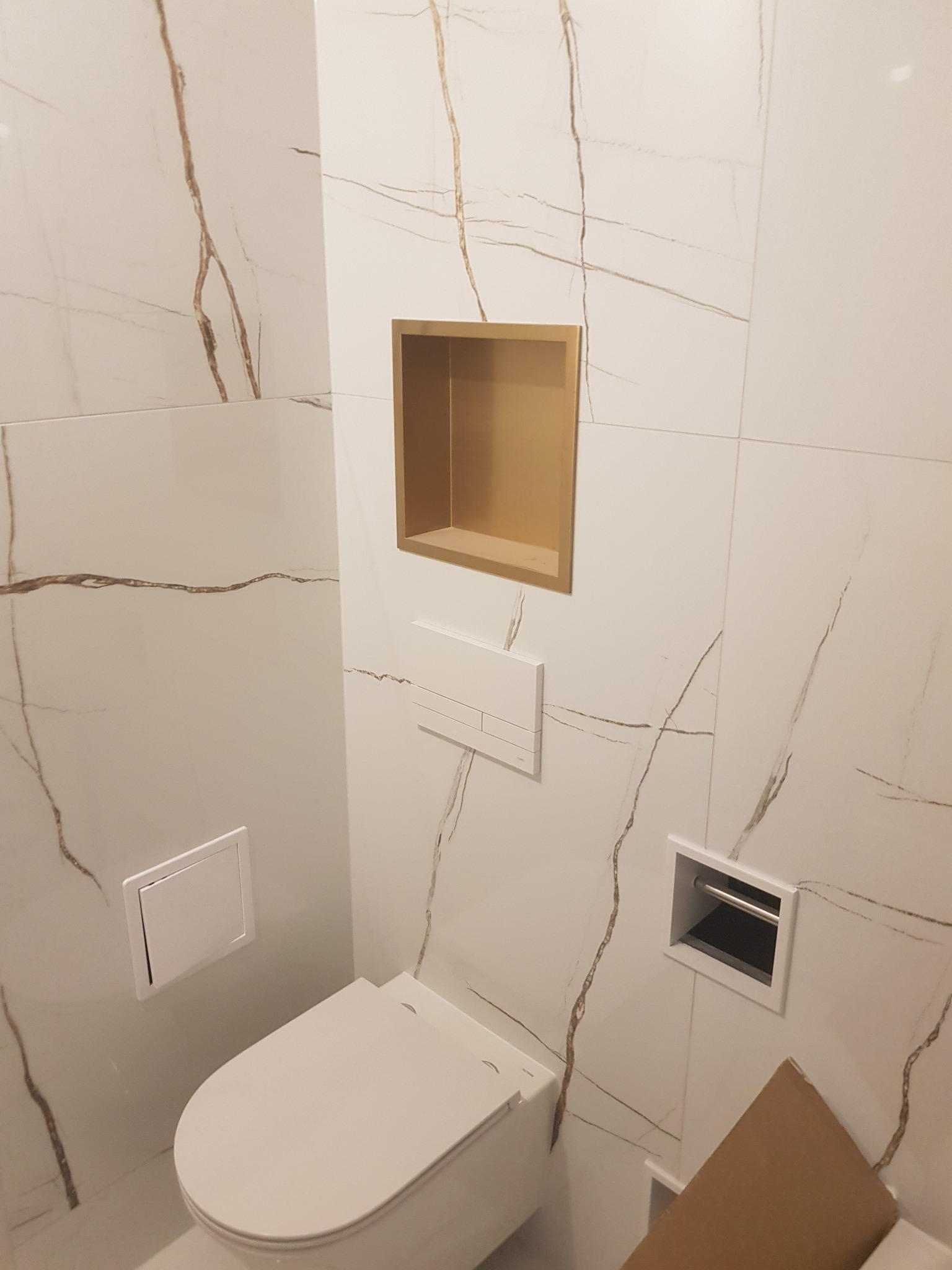 Nowoczesna łazienka z białą ceramiką i płytkami imitującymi marmur z brązowymi żyłami, wnęka w ścianie wykończona złotym kolorem, widoczny fragment kartonu na podłodze.