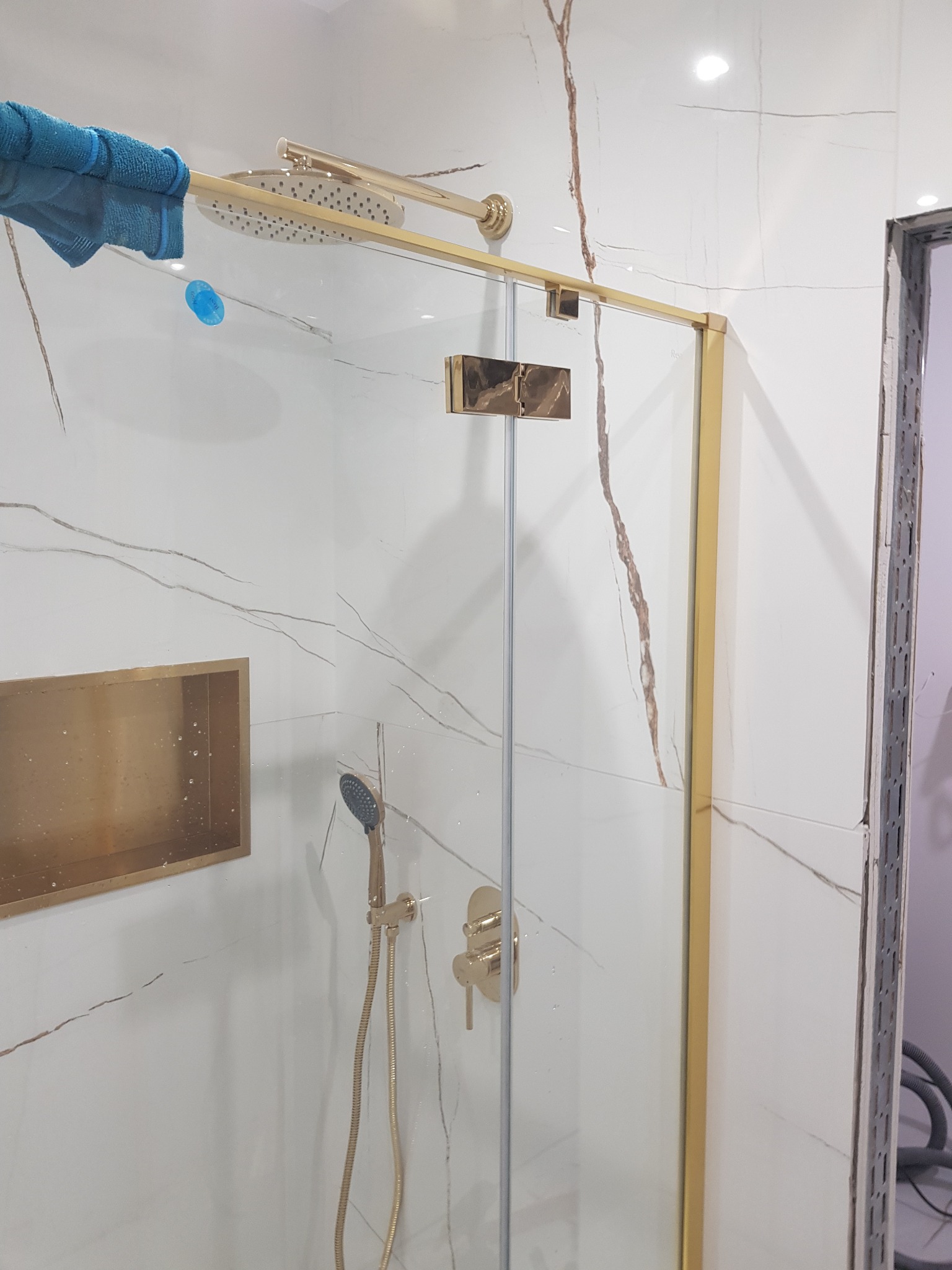 Nowoczesna kabina prysznicowa z złotymi elementami i białymi płytkami imitującymi marmur, widoczna niebieska ściereczka na górnej ramie, w tle przygotowane miejsce do wykończenia.