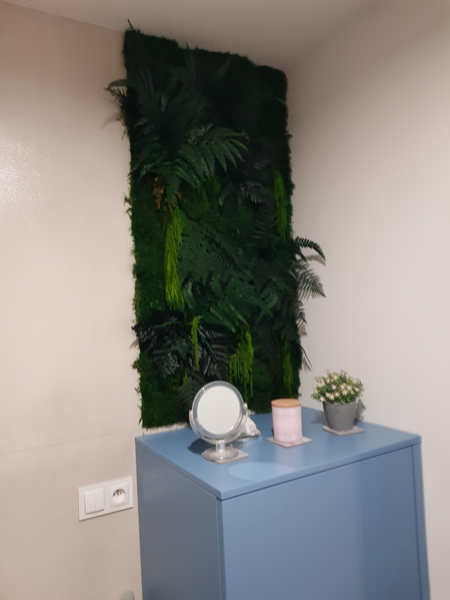 Ściana z mchu i paproci w pomieszczeniu, niebieska szafka z lustrem i kwiatami w doniczce, gniazdko elektryczne na ścianie.