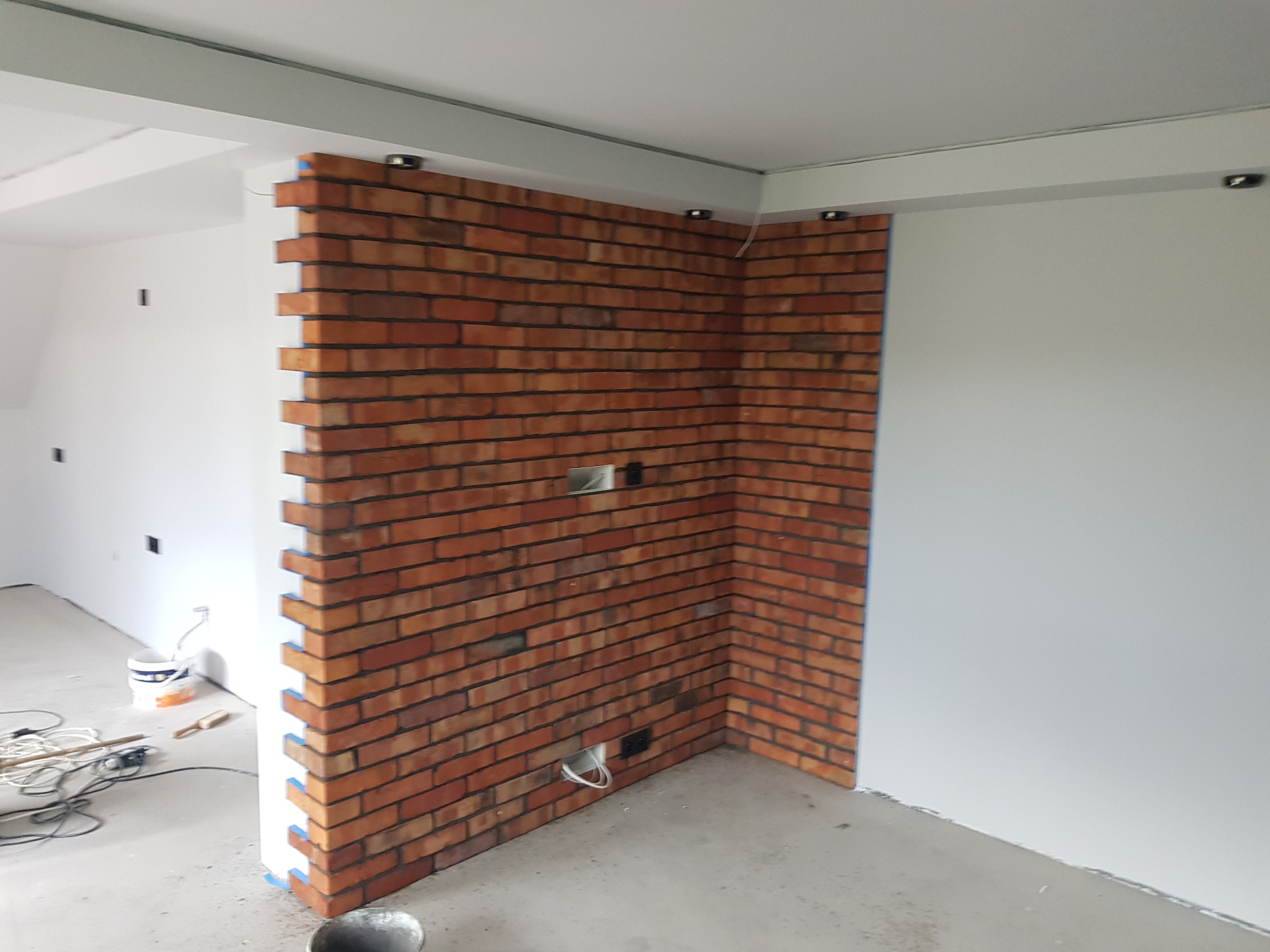 Fragment ściany z cegły klinkierowej w trakcie prac wykończeniowych, widoczne puszki elektryczne, resztki zaprawy na podłodze i taśma malarska zabezpieczająca narożnik.