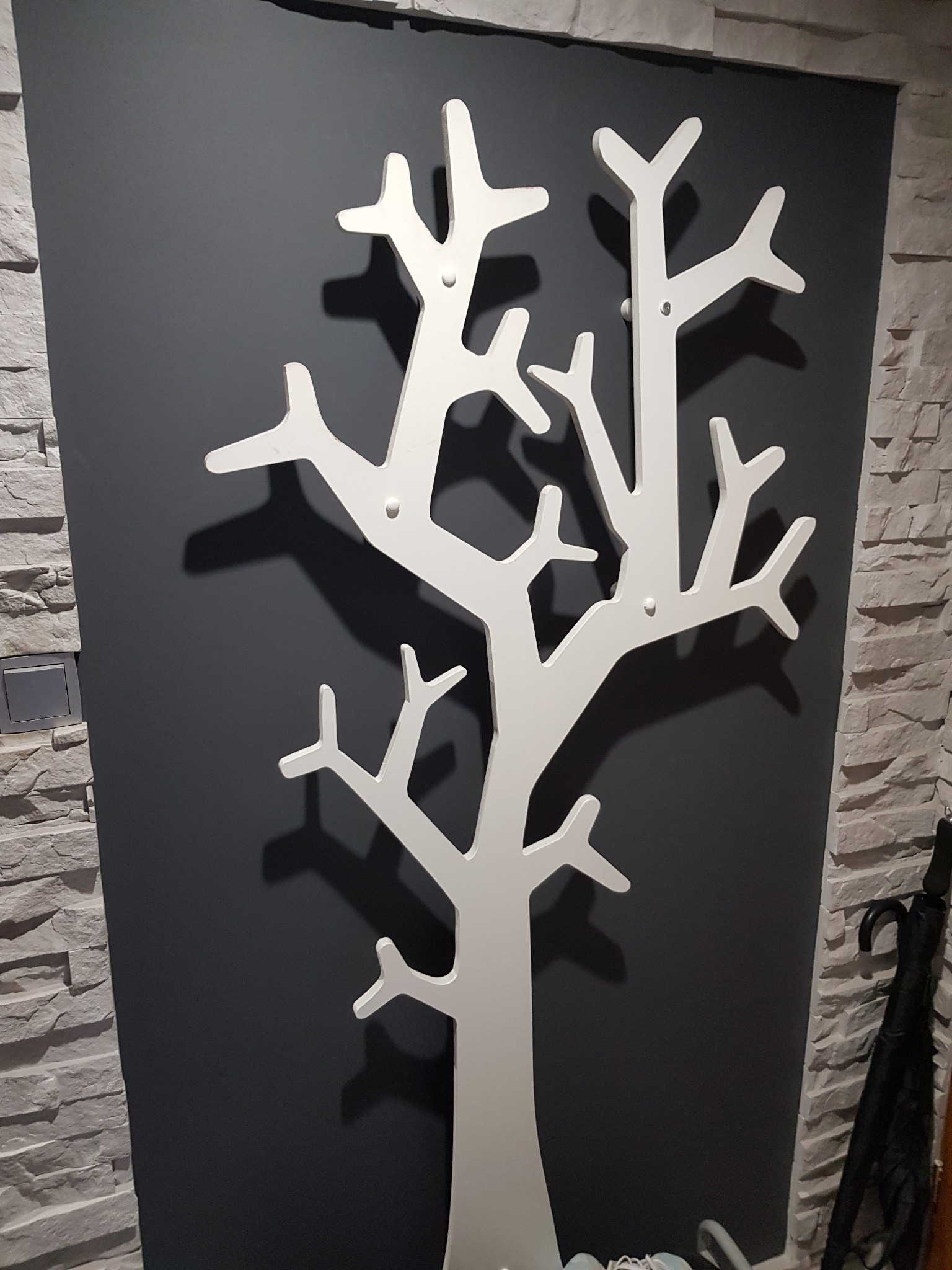 Biały, dekoracyjny wieszak w kształcie drzewa na szarej ścianie, z fragmentem ściany z kamienia dekoracyjnego po bokach.