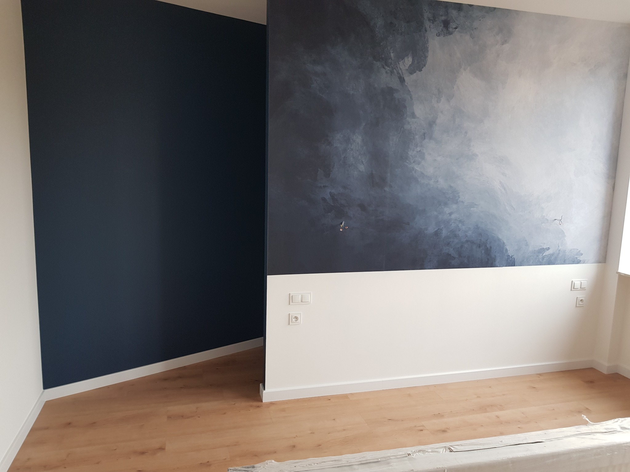 Pokój po odświeżeniu: ściana w kolorze głębokiego granatu sąsiaduje z białą ścianą, nad którą znajduje się dekoracyjna malatura w odcieniach szarości i błękitu, przypominająca morski pejzaż. Jasne...