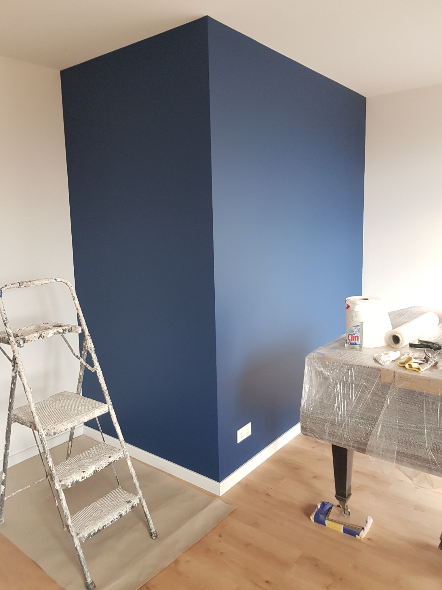 Pomieszczenie po malowaniu z granatową ścianą narożną, białe ściany boczne, drabina malarska z resztkami farby, stół zabezpieczony folią i akcesoria malarskie.